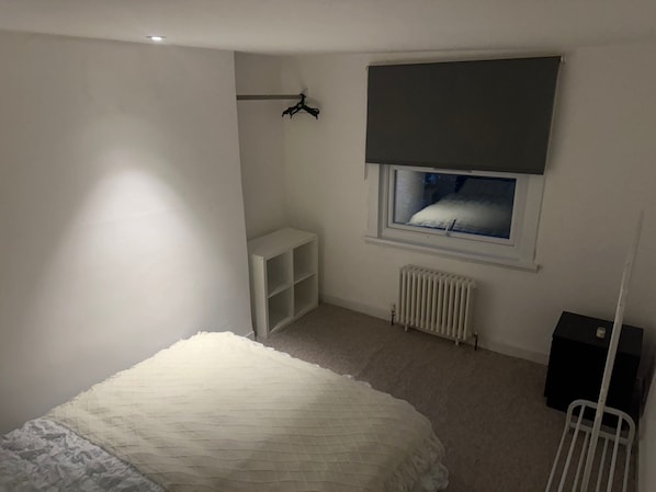 Ein Schlafzimmer mit Bett, Heizkörper, Fenster mit Jalousie, weißem Kleiderschrank und Kleiderhaken.