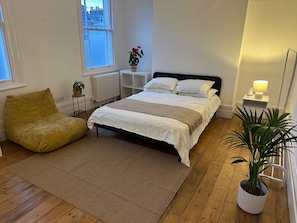 Un dormitorio con cama, una silla, un radiador, una ventana y una planta.