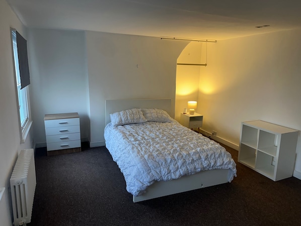 A bedroom with a bed, a nightstand with a lamp, a dresser, and a shelving unit.