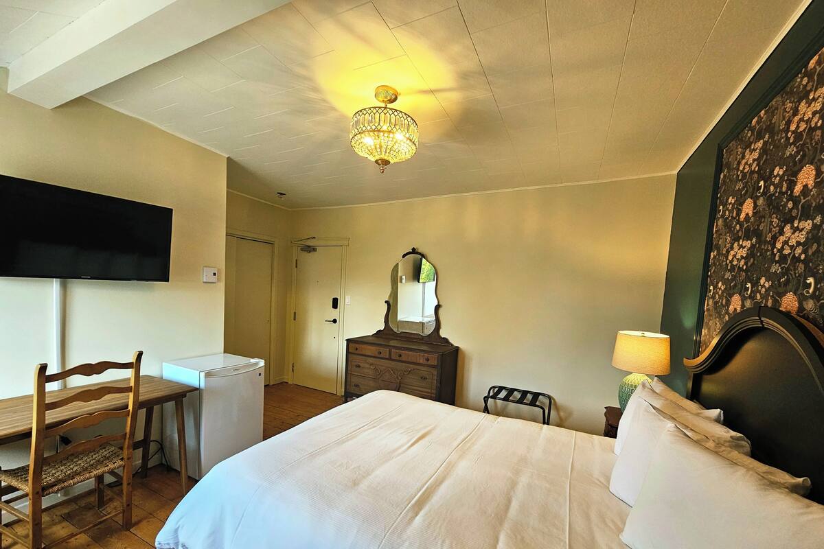 A bedroom with a bed, a dresser, a chair, a desk, a television, and a chandelier.