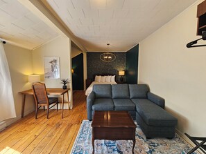 Um espaço de estar compacto com piso de madeira, um sofá cinza, uma mesa pequena, uma cadeira e uma cama com cabeceira escura.