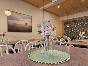 Uma sala de jantar com mesas redondas, cadeiras, um vaso com flores, um quadro na parede e um papel de parede estampado.