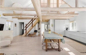 Ein geräumiges Zimmer mit hoher Decke, freiliegenden Holzbalken, einem Esstisch mit Stühlen, einer Treppe und einer Küchenzeile.