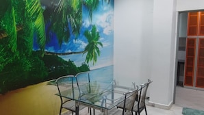 Ein Zimmer mit Deckenventilator, einem Glastisch mit Stühlen, einem Wandbild mit Strandmotiv und einer Tür mit orangefarbenen Paneelen.
