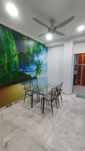 Ein Zimmer mit Deckenventilator, einem Glastisch mit Stühlen, einem Wandbild mit Strandmotiv und einer Tür mit orangefarbenen Paneelen.