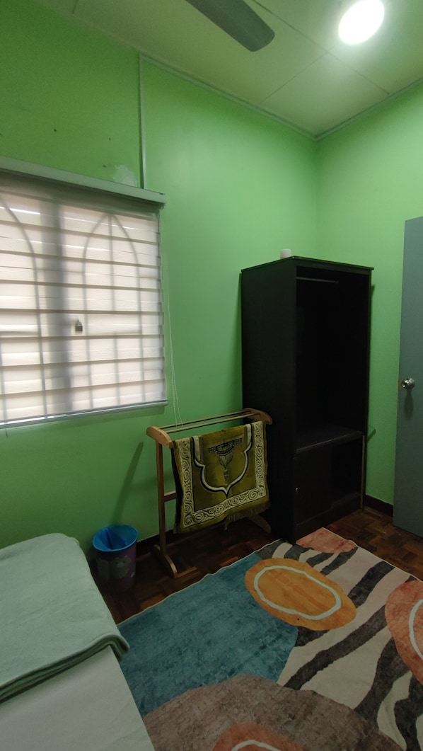 A room with a green wall, a window with blinds, a black wardrobe, a small table with a towel, and a patterned rug.