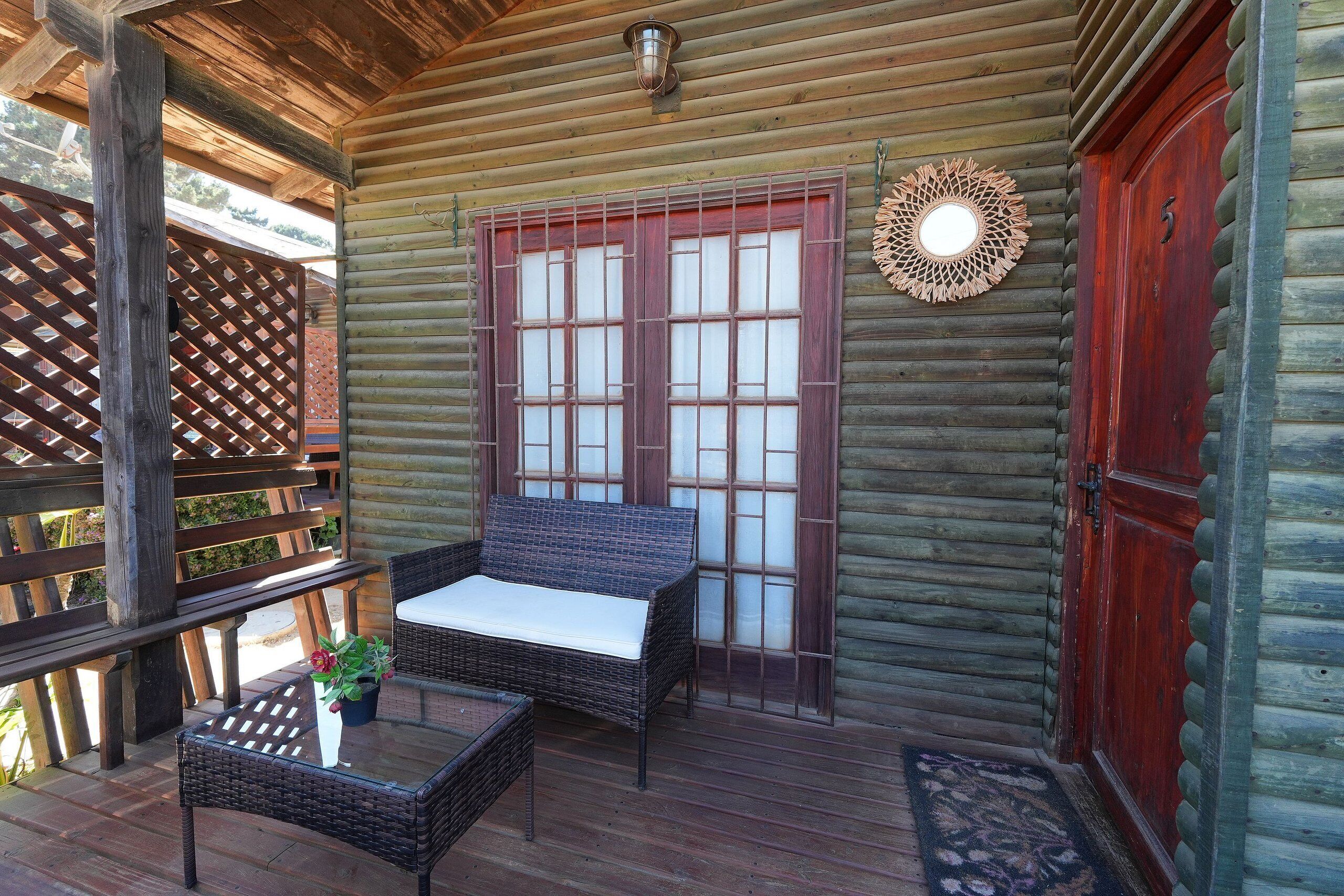Een vakantiehuis van hout met een houten terras, een rieten bank, een tafel en een decoratieve spiegel.