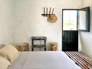 A bedroom with a bed, a small table, a chair, a round stool, a hat on the wall, and a door with a mirror. - Cortijo with eclectic décor near Almería (Albaricoques)