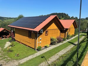 A wooden cabin with a red roof, solar panels, and a paved pathway leading to a picnic area. - Komfortowy domek nad Soliną z widokiem. Taras, grill, zwierzęta mile widziane! (Zawóz)