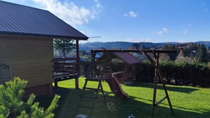 A wooden play structure in a backyard with a house and a view of distant hills.