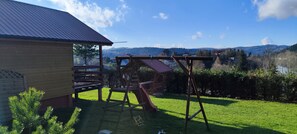 A wooden play structure in a backyard with a house and a view of distant hills. - Komfortowy domek nad Soliną z widokiem. Taras, grill, zwierzęta mile widziane! (Zawóz)