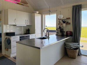 Une cuisine moderne avec des meubles blancs, un Ăźlot central dotĂ© dâun Ă©vier, dâune machine Ă cafĂ© et offrant une vue sur un champ verdoyant par la fenĂȘtre.