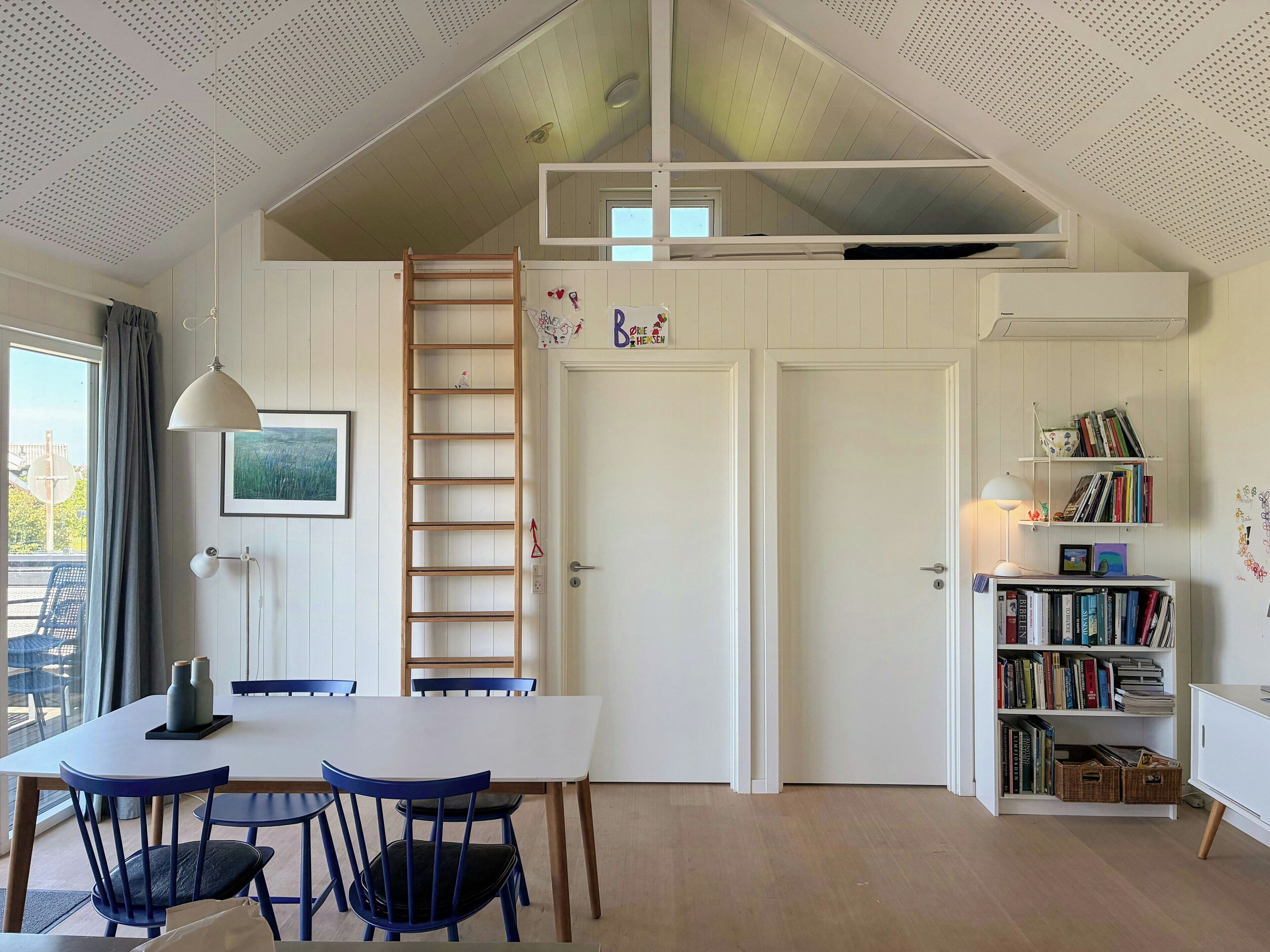 Um quarto com porta branca, estante de livros, mesa de jantar com cadeiras, escada e luminária pendente.