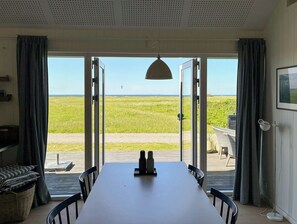 Une piĂšce dotĂ©e dâune grande table, de chaises, dâun luminaire suspendu et offrant une vue sur un champ verdoyant grĂące Ă des portes ouvertes.