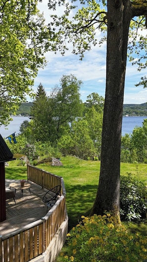 Una terraza de madera con mobiliario exterior, un árbol grande y vistas a un lago y colinas.