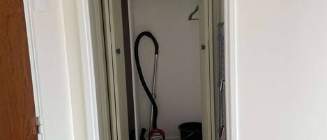 A hallway leading to a closet with a vacuum cleaner and a bucket.