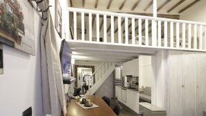 Um quarto estilo loft com teto de madeira, área de jantar com mesa e cadeiras, cozinha com armários brancos e escada para um andar superior.