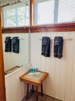 Ein Badezimmer mit weißem Holzvertäfelung, Holzbalken, einem Spiegel, Handtüchern und Pflegeprodukten auf einem Holztisch.