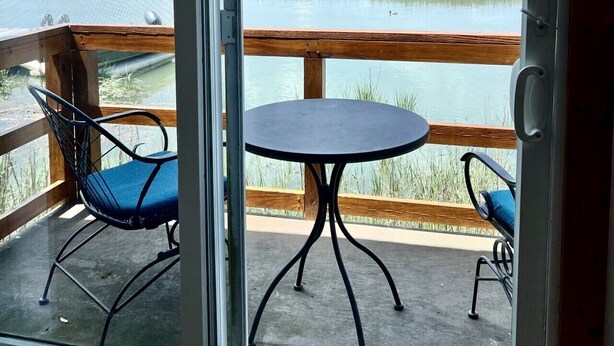 Ein Zimmer mit einer Glastür zum Balkon, auf dem sich ein Tisch und zwei Stühle befinden, mit Blick auf ein Gewässer und eine entfernte Landschaft.