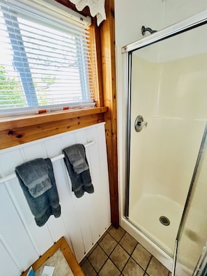 A bathroom with a shower, a window with blinds, and two towels hanging on a rack.