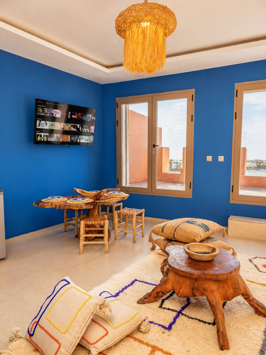 A room with a blue wall, a TV mounted on it, a dining table with chairs, a hanging light fixture, and a window with a view.
