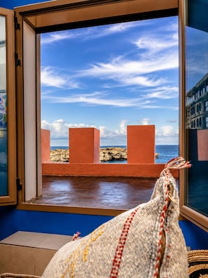 Ein Zimmer mit Blick auf eine felsige Küste durch ein Fenster, eine gewebte Tasche auf dem Boden und eine blaue Wand.