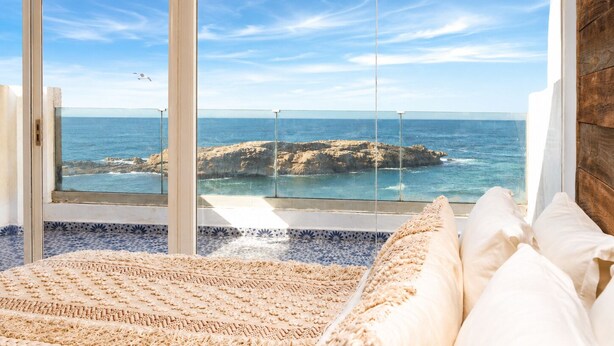 Une chambre avec une grande fenêtre donnant sur la mer, un lit avec une literie blanche et un tapis à motifs.