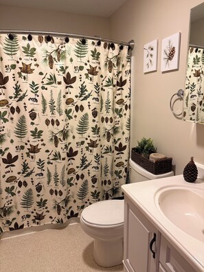 Une salle de bain avec un rideau de douche orné de motifs végétaux, des toilettes, un lavabo avec un miroir et une petite plante posée sur le comptoir.