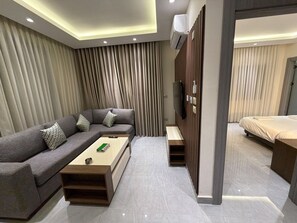 A modern hotel room with a sectional sofa, a wooden coffee table, a TV mounted on a wooden panel, and a bed with a white bedspread. - 104 JRE Apartment With Stunning Boulevar (Amman)