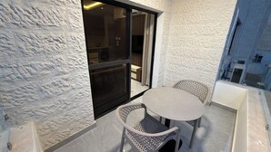 En balkong med en vit, strukturerad vägg, en skjutdörr i glas som leder in till ett rum, och två flätade fåtöljer runt ett litet runt bord.