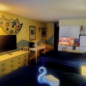 Ein Hotelzimmer mit zwei Betten, einem Schreibtisch, einem Sessel, einem Schminktisch, einem Fernseher und einer Küchenzeile.