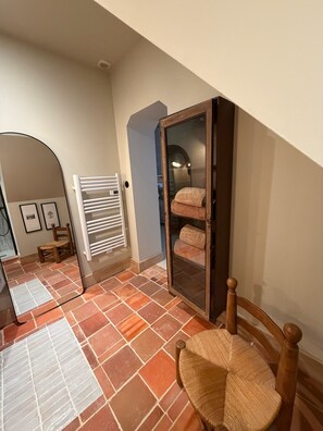 A room with a tiled floor, a wooden chair, a white radiator, a glass cabinet with towels, and a mirror on the wall. - Vrbo Property (Chinon)