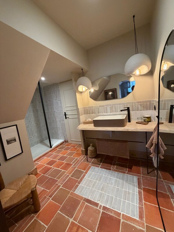A modern bathroom with a sloped ceiling, a large mirror, a vanity with a sink, and a shower area. - Vrbo Property (Chinon)