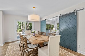 A dining area with a large wooden table, wicker chairs, a hanging light fixture, and a sliding barn door. - Seabrook Exclusives | 2963 Seabrook Island Rd (Seabrook Island)