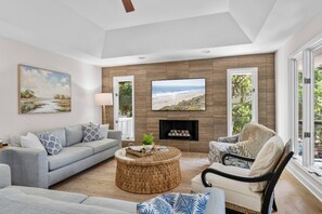 A living room with a sofa, armchair, coffee table, fireplace, and large windows. - Seabrook Exclusives | 2963 Seabrook Island Rd (Seabrook Island)