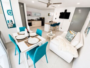Ein modernes Wohnzimmer mit einem weißen Sofa, einem Essbereich mit Tisch für vier Personen und einer Küche im Hintergrund.