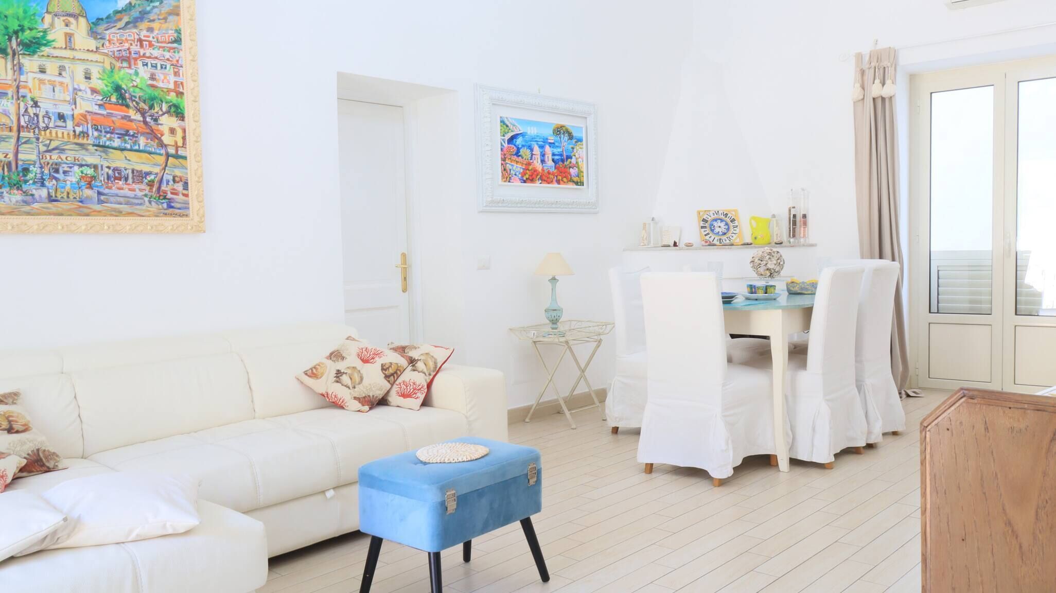 A living room with a white sofa, a blue ottoman, a dining table with chairs, and a painting on the wall.