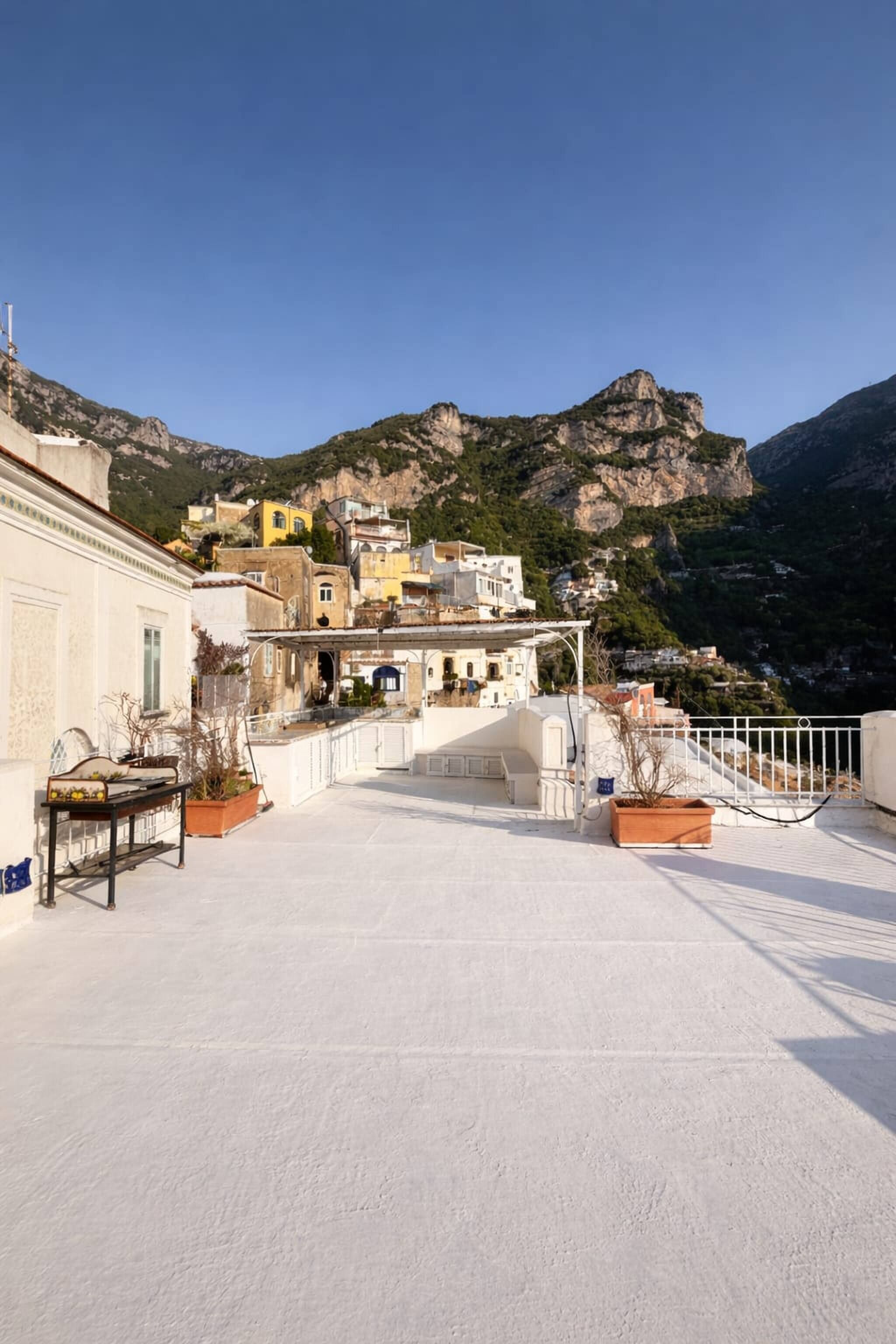 A rooftop terrace with a view of a mountainous landscape, featuring buildings with balconies and potted plants.