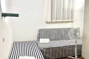 Ein Zimmer mit Bett, einem kleinen Tisch, einem Ventilator und einem Fenster mit leichtem Vorhang.
