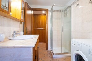 A bathroom with a sink, mirror, wooden cabinets, a glass shower enclosure, and a washing machine. - Laide House - Walking Beach + Garage + A/C (Castelsardo)
