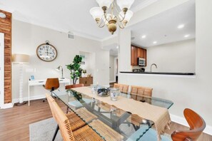 A modern dining area with a glass table, wooden chairs, a chandelier, a clock, and a kitchen in the background. - Reunion Resort 3 Bed, 2 Bath with Pool Views & Golf Course (Kissimmee)