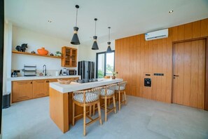 A modern kitchen with wooden cabinets, a central island with bar stools, pendant lighting, and an air conditioning unit. - K Club Cemagi Villa 1 (Badung)