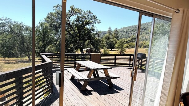 A wooden deck with a picnic table and chairs, a glass door leading outside, and a view of a forested area. - 3-bedroom house in peaceful Sierras on 4.5 acres along road to Bass Lake (Wishon)