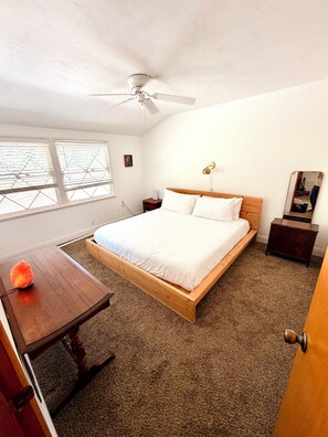 A bedroom with a bed, a ceiling fan, a nightstand, a bedside table, a heater, and a window with blinds.