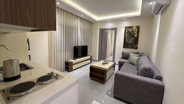 A modern living room with a flat-screen TV, a sofa, a coffee table, and a kitchen area with a kettle and a sink. - 105 JRE Apartment With Stunning Boulevar (Amman)