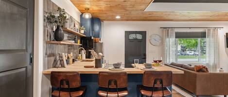 A modern kitchen with a wooden ceiling, a bar area with stools, a built-in oven, and a large window with a view of the outside.