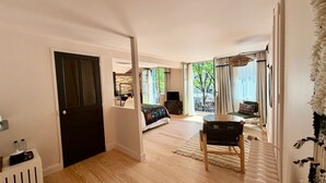 Een ruime woonkamer met houten vloer, een eettafel met stoelen, een bank en een groot raam met uitzicht op bomen.