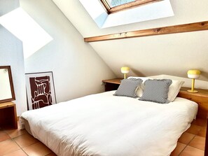 Ein Schlafzimmer mit schrägen Wänden, einem Bett mit weißer Bettwäsche, zwei Nachttischlampen und einem gerahmten Kunstwerk an der Wand.