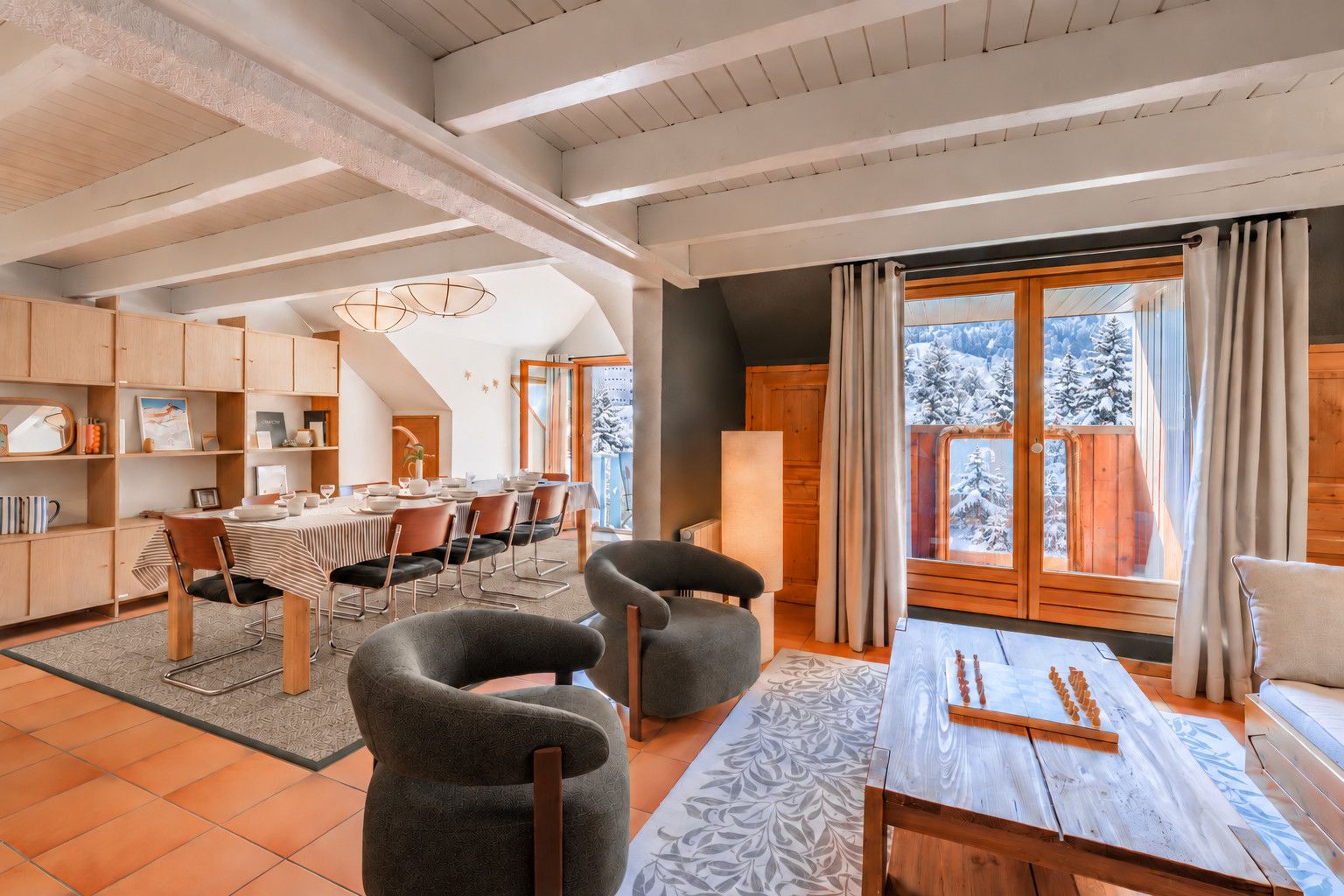 A spacious living area with a dining table, two armchairs, and a view of snowy mountains through large windows.