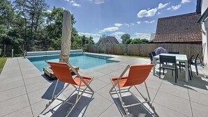 Een achtertuin met een zwembad, twee oranje ligstoelen, een overdekte patio tafel en een houten omheining.